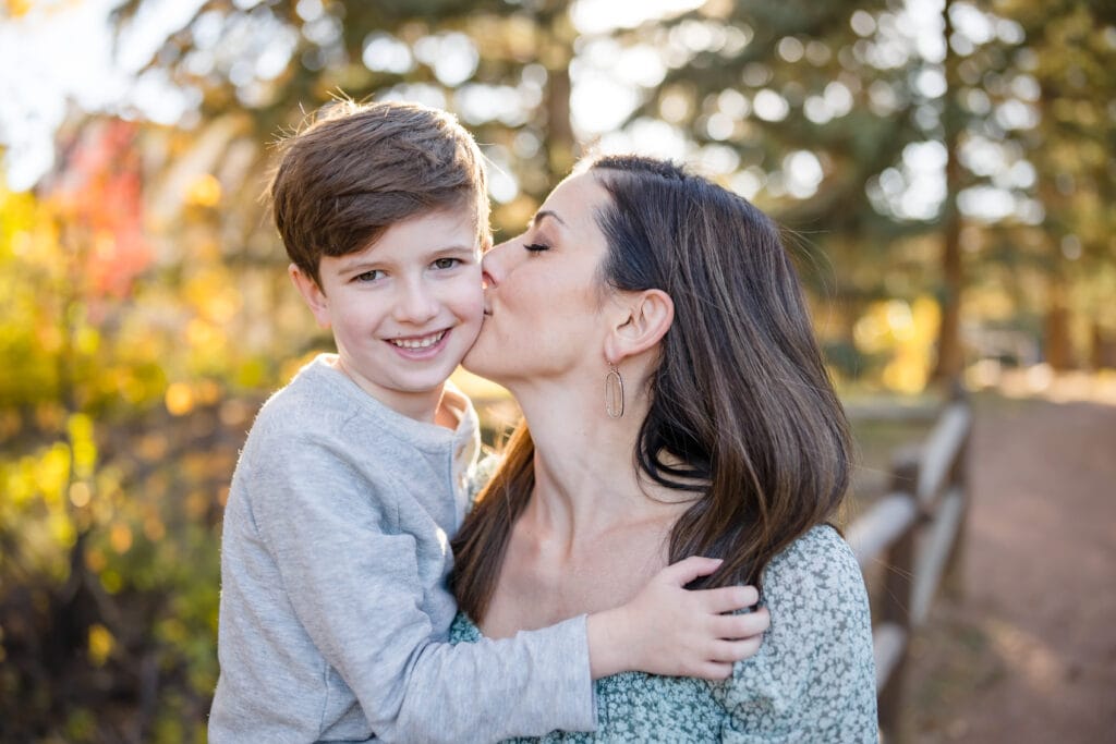Mom kissing son on the cheek with setting sun filtered through trees in the background
