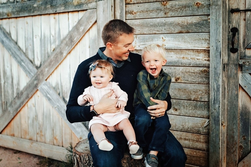 Dad seated in front of barn holding laughing toddlers