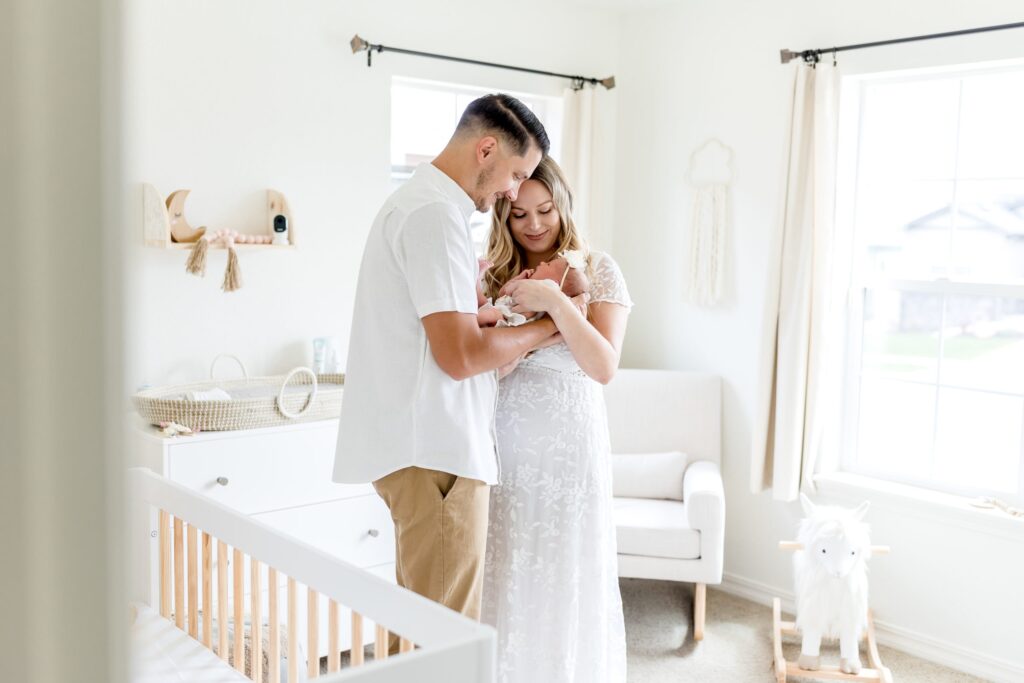 Husband and wife cuddle close together during their in home newborn photoshoot, holding their newborn in their light, airy, neutral nursery just a few days after they brought their baby home from the hospital
