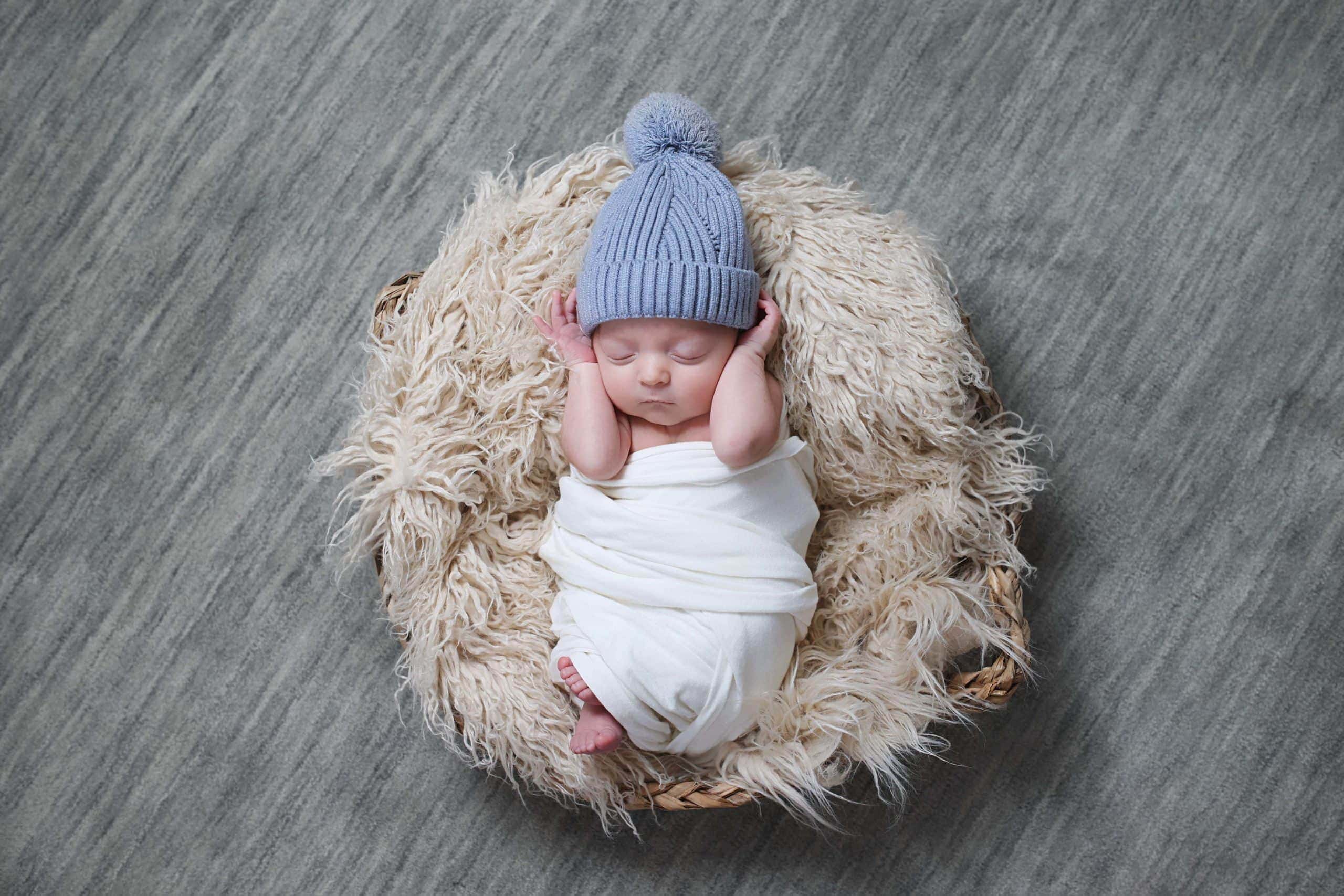 Studio style newborn image of a baby wrapped in a white swaddle, posed in a curved bowl with a faux fur blanket background and blue knitted hat