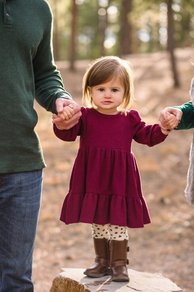 Toddler girl in maroon dress and boots holding on to mom and dad's hands