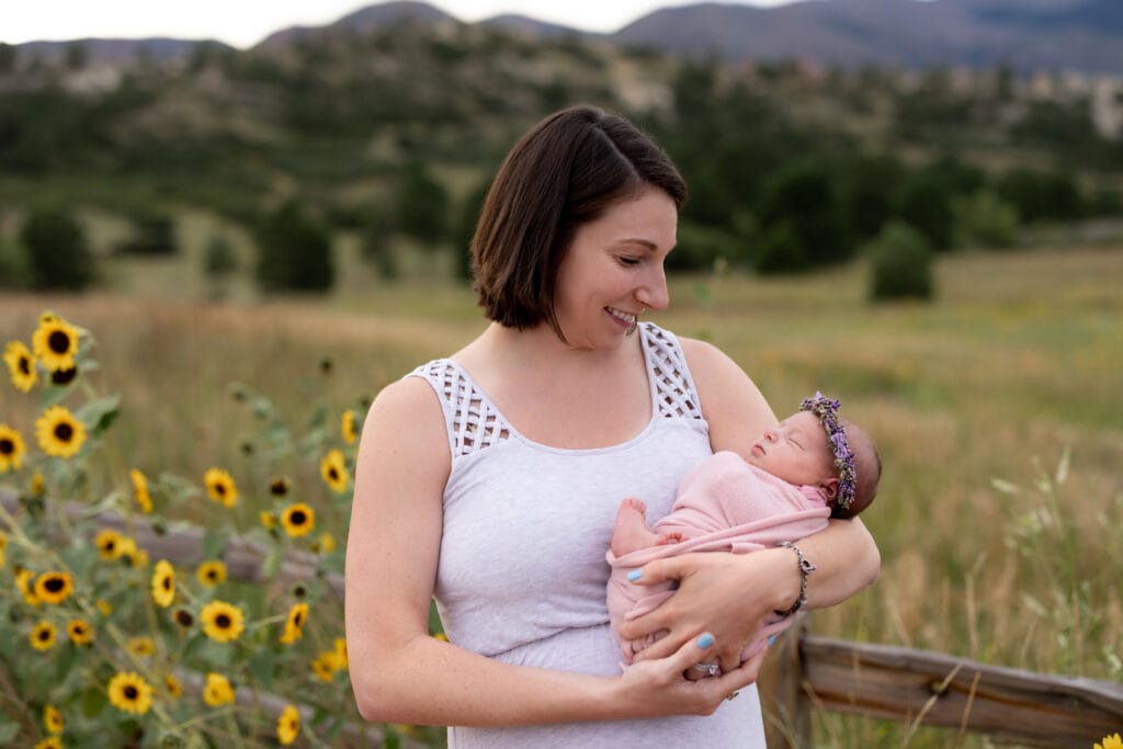 Mother standing by fence and smiling down at baby daughter during outdoor newborn session in Ute Valley Park, Colorado