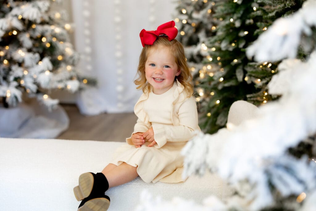 Toddler girl in white dress and red bow smiling for Christmas card photos in Colorado Springs studio