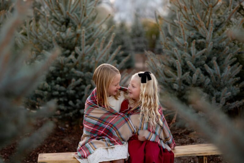 Sisters wrapped in plaid blanket and seated on bench with evergreen trees in the background