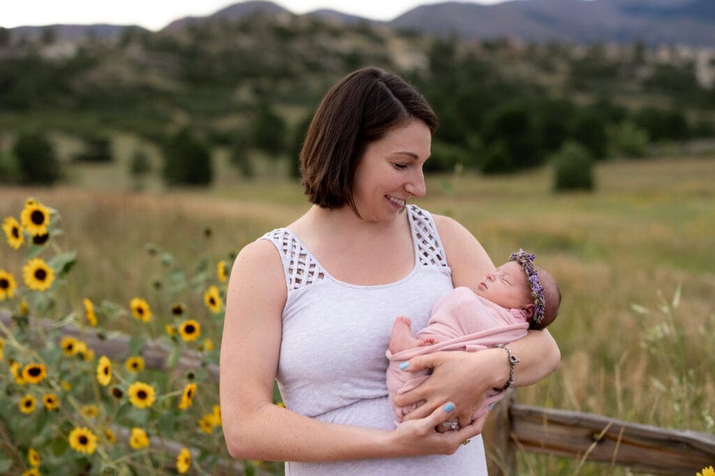 Woman holds her newborn near a rustic wooden fence overgrown with wild sunflowers during summer family photos in Colorado Springs