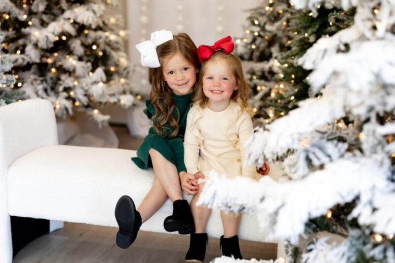 Sisters seated on white bench by Christmas trees during holiday photoshoot in Colorado