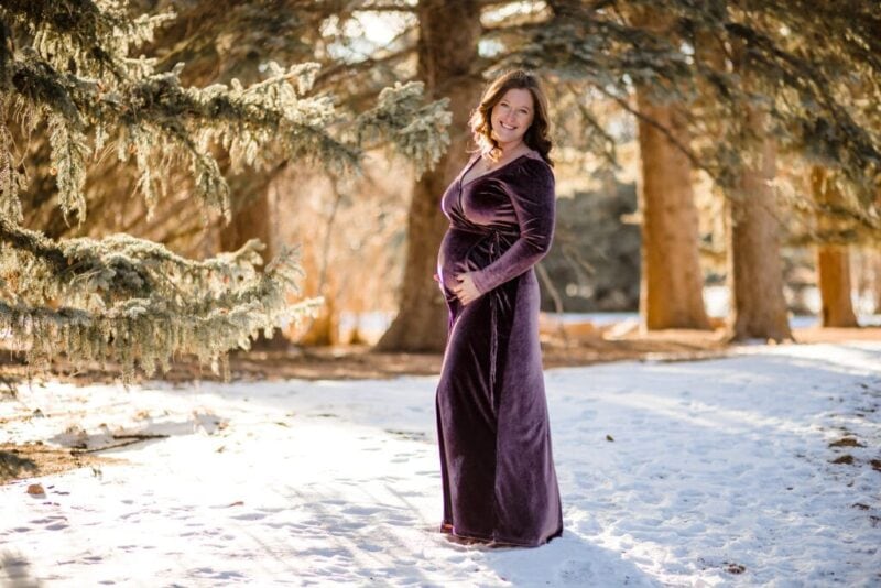 woman in purple maternity gown standing in snow with evergreen trees in the background