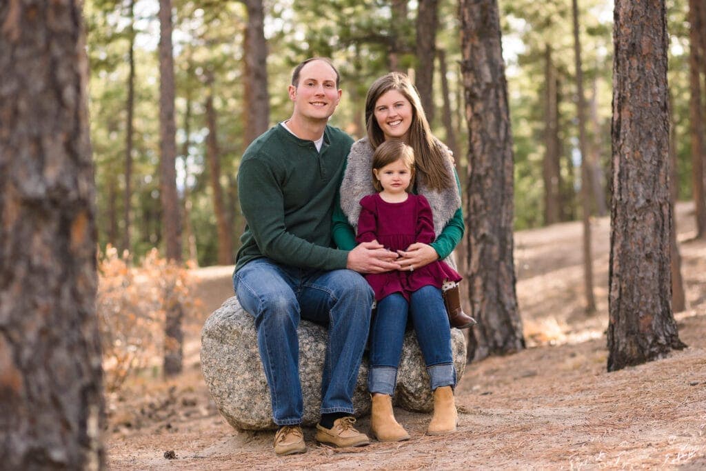Parents sitting on stone in woods with toddler daughter on their lap