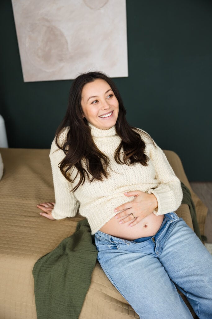 Pregnant woman in jeans and sweater seated on bed in lifestyle studio in Colorado Springs