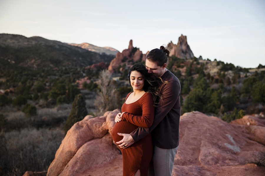 Husband embraces pregnant wife from behind as they stand in Garden of the Gods with rugged rock formations in the backdrop