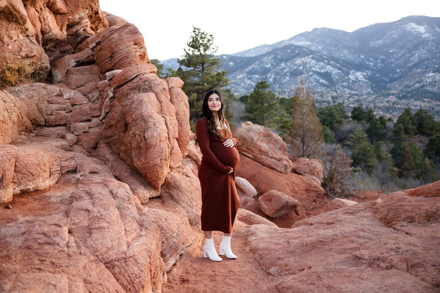 Woman in rust-colored maternity gown and white boots gazes up at cliffs in Colorado Springs with mountains in the background